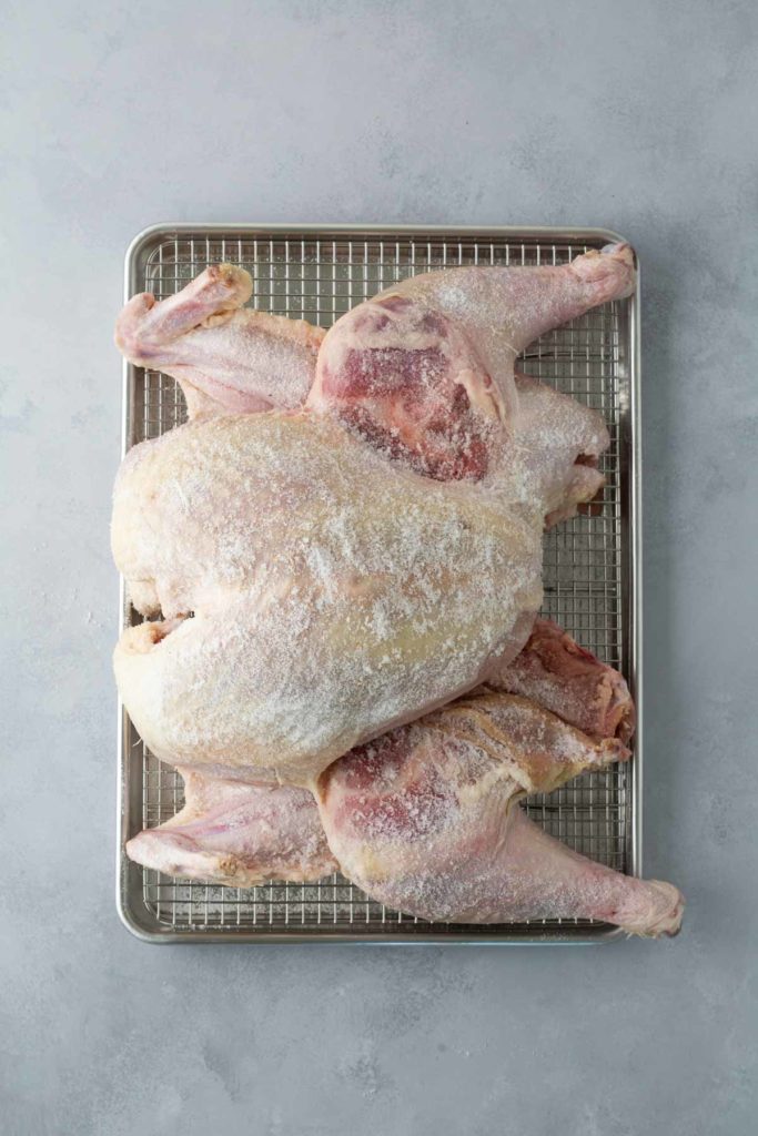 A raw, spatchcocked chicken is coated with salt and placed on a metal rack over a tray against a gray background.