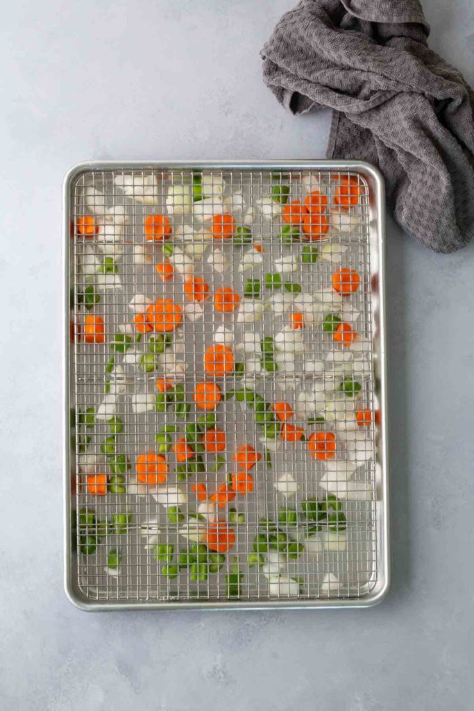 Chopped carrots, onions, and green peppers on a wire rack over a baking sheet, with a gray cloth beside it.