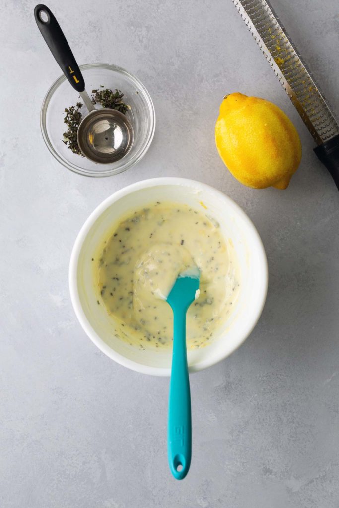 A bowl of creamy sauce with herbs, a blue spatula, a lemon, a small bowl with herbs, and a grater on a gray surface.