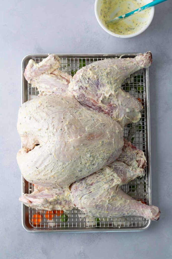 A raw spatchcocked turkey is placed on a wire rack over a baking sheet, coated with a creamy marinade. A bowl with marinade and a brush is nearby.