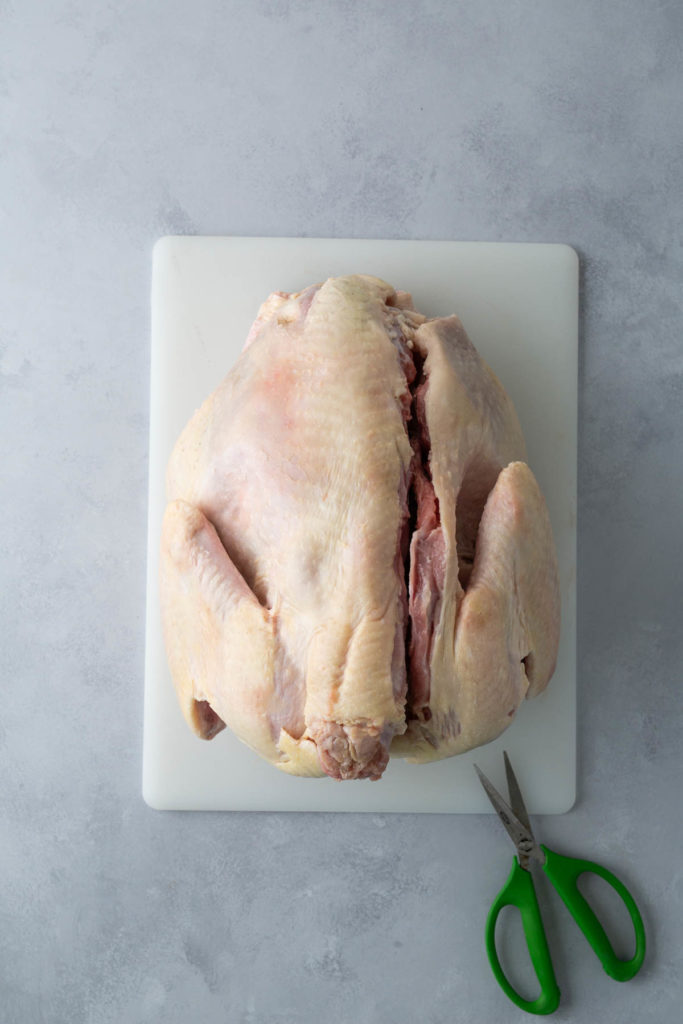 Raw whole chicken on a white cutting board with green-handled scissors beside it, on a gray textured surface.