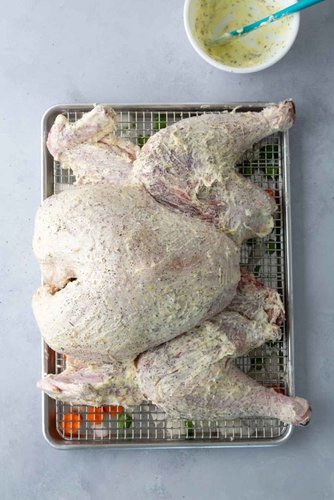 Raw spatchcocked turkey seasoned with a creamy herb mixture on a wire rack over a baking sheet, with a bowl of extra seasoning nearby.