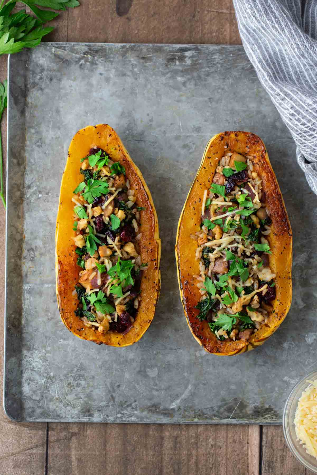 Two halved squash on a baking sheet, filled with a mixture of vegetables, nuts, and herbs, garnished with fresh parsley.