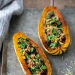 Two roasted delicata squash halves filled with a mixture of greens, nuts, and herbs, placed on a metal tray with a striped cloth beside it.