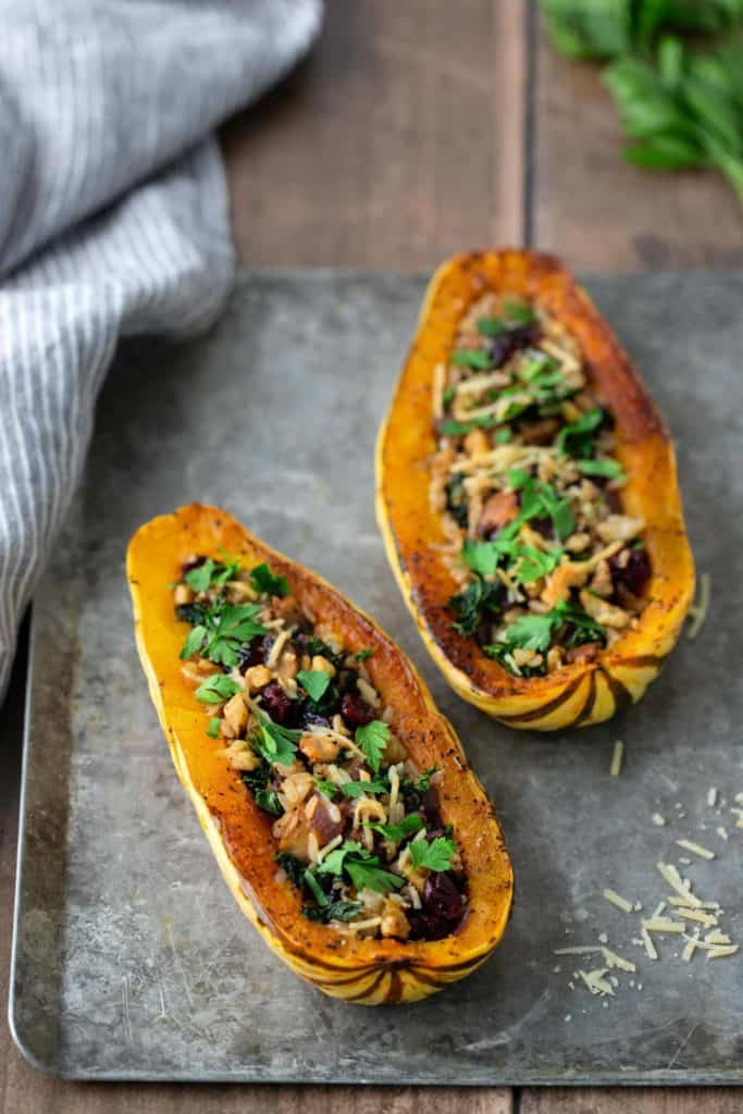 Two roasted delicata squash halves filled with a mixture of greens, nuts, and herbs, placed on a metal tray with a striped cloth beside it.