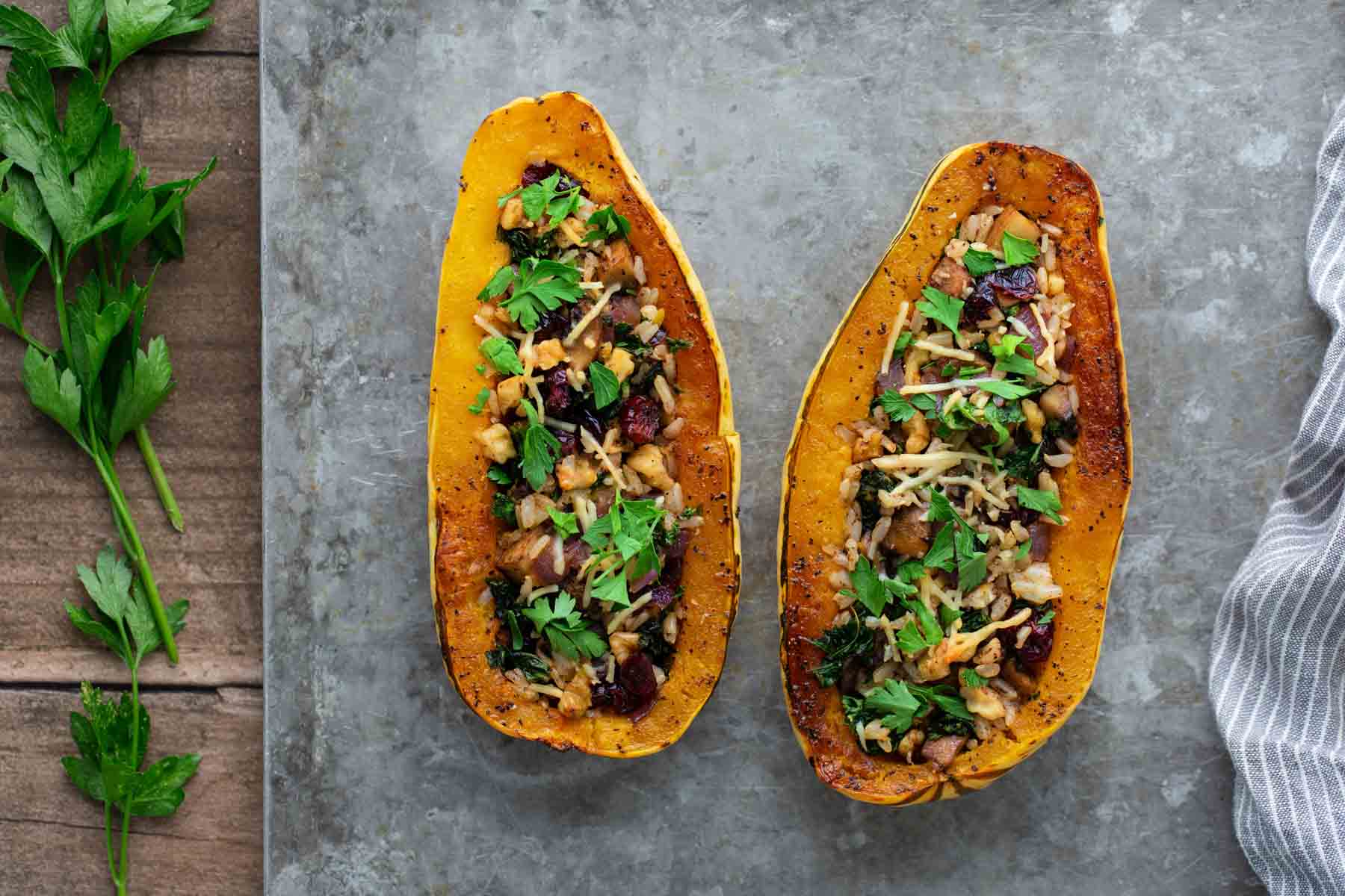 Two roasted squash halves filled with a mixture of greens, nuts, and cheese on a textured surface, with parsley leaves on the side.