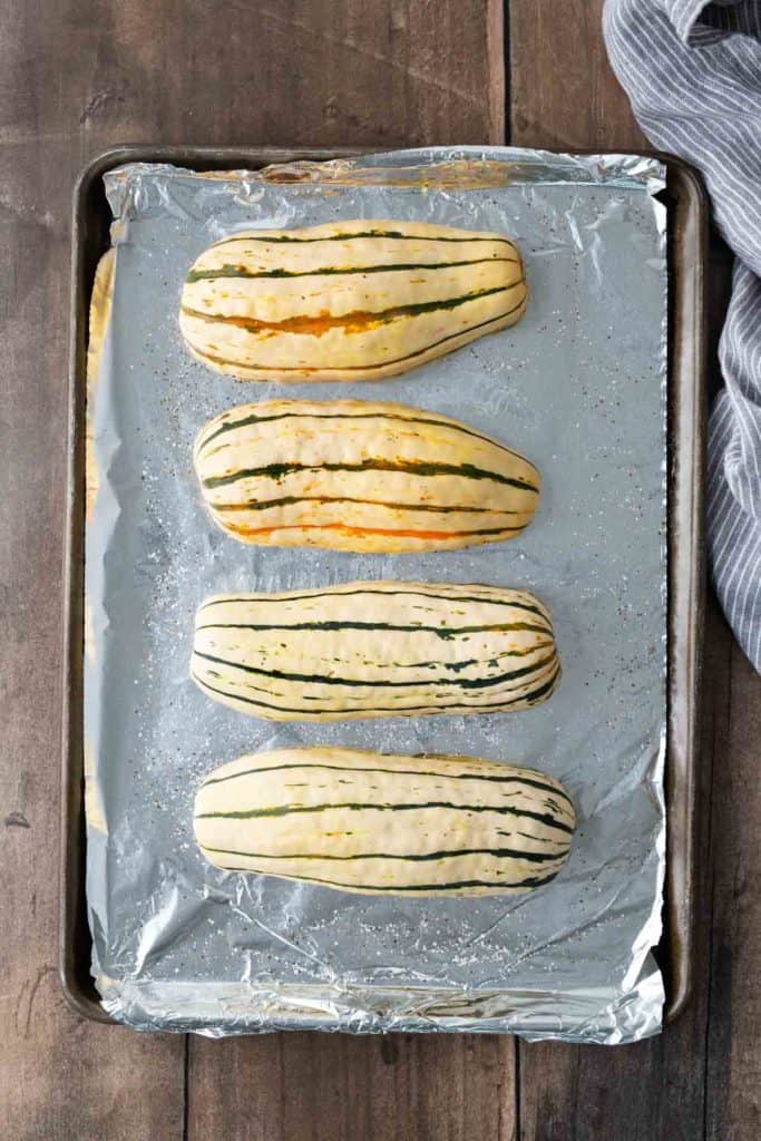 Four uncooked striped squash lined up on foil-covered baking sheet, wooden surface below.