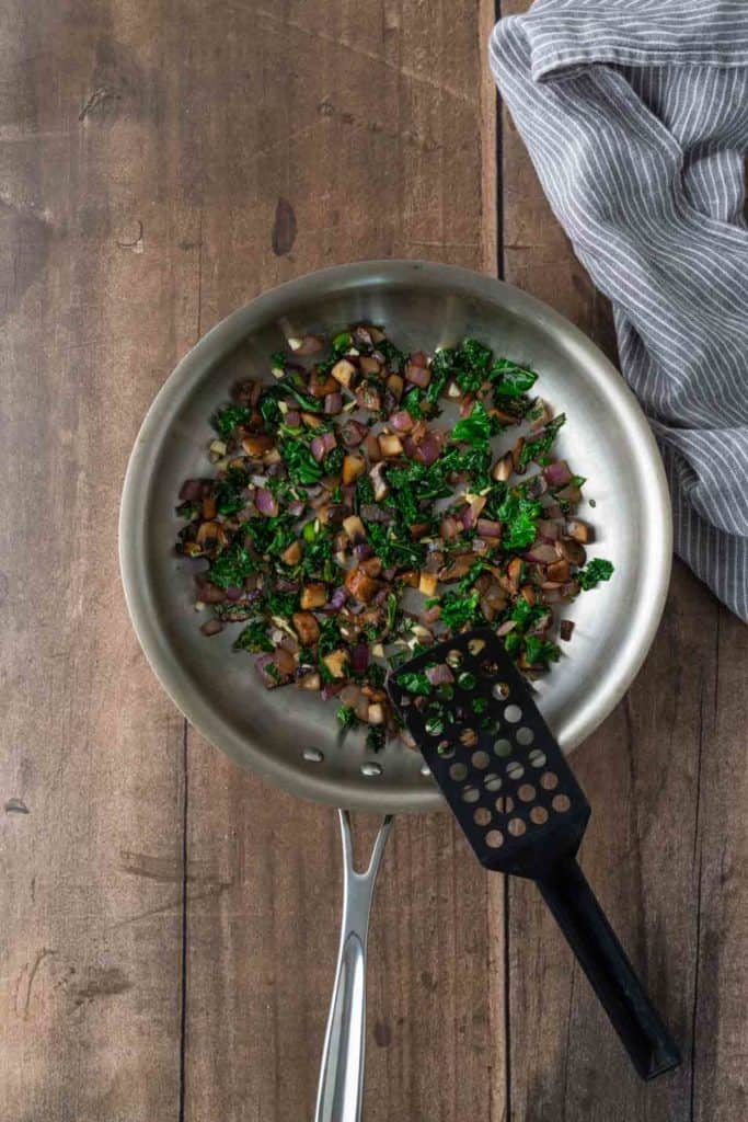 A pan with sautéed vegetables, including kale and onions, on a wooden surface next to a striped cloth. A black spatula rests in the pan.