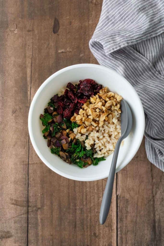 A white bowl with brown rice, chopped walnuts, cranberries, mushrooms, and greens, placed on a wooden surface with a striped cloth beside it. A gray spoon is on the bowl's edge.