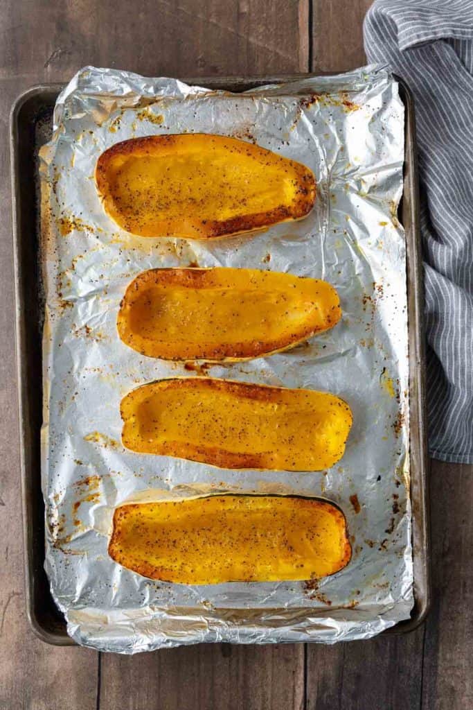 Four halves of roasted butternut squash on a foil-lined baking sheet, seasoned with salt and pepper.