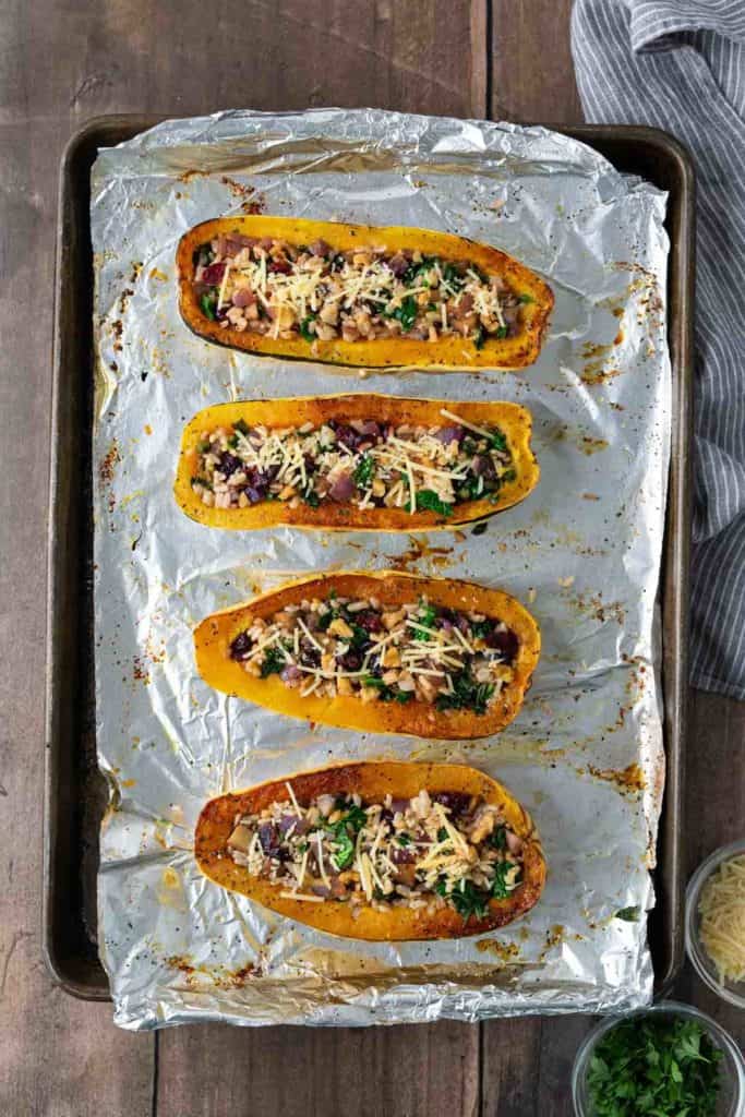 Four stuffed squash halves with toppings on a foil-lined baking sheet, garnished with cheese and herbs.