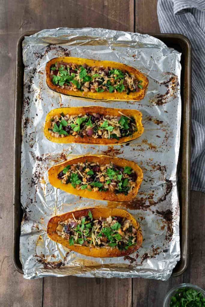 Four stuffed squash halves on a foil-lined baking sheet, topped with herbs and vegetables.