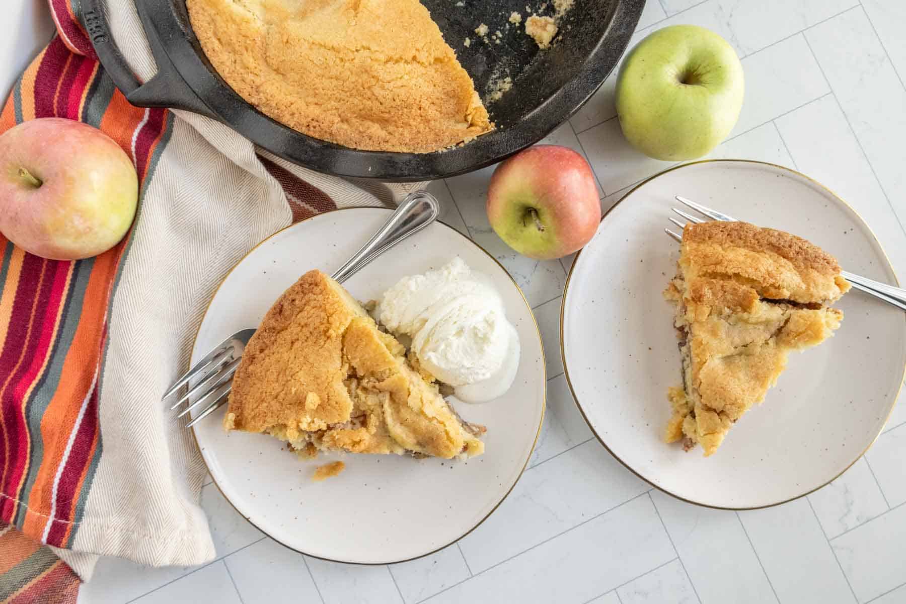 Two plates with apple pie slices, one with ice cream, next to whole apples and a striped cloth. A cast iron pan with remaining pie is in the background.