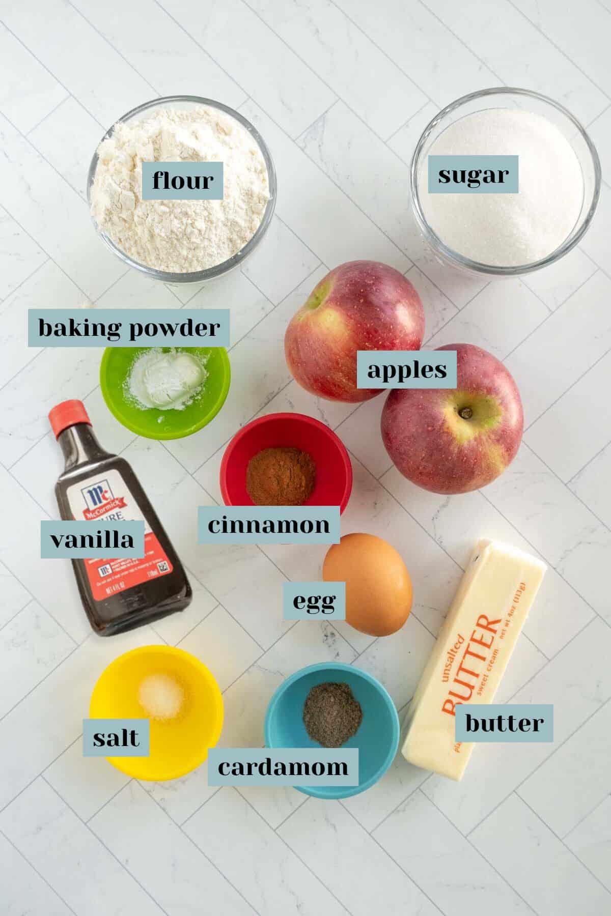 Ingredients for a recipe are laid out on a white surface. Items include flour, sugar, baking powder, apples, vanilla, cinnamon, an egg, butter, salt, and cardamom.