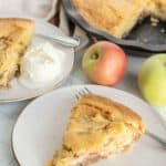 Slice of apple pie on a plate with a fork, served with a scoop of vanilla ice cream. Whole pie and apples in the background.