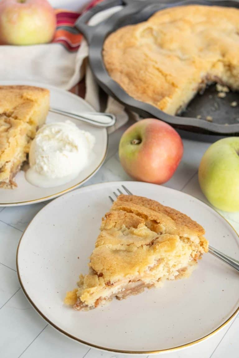 Slice of apple pie on a plate with a fork, served with a scoop of vanilla ice cream. Whole pie and apples in the background.