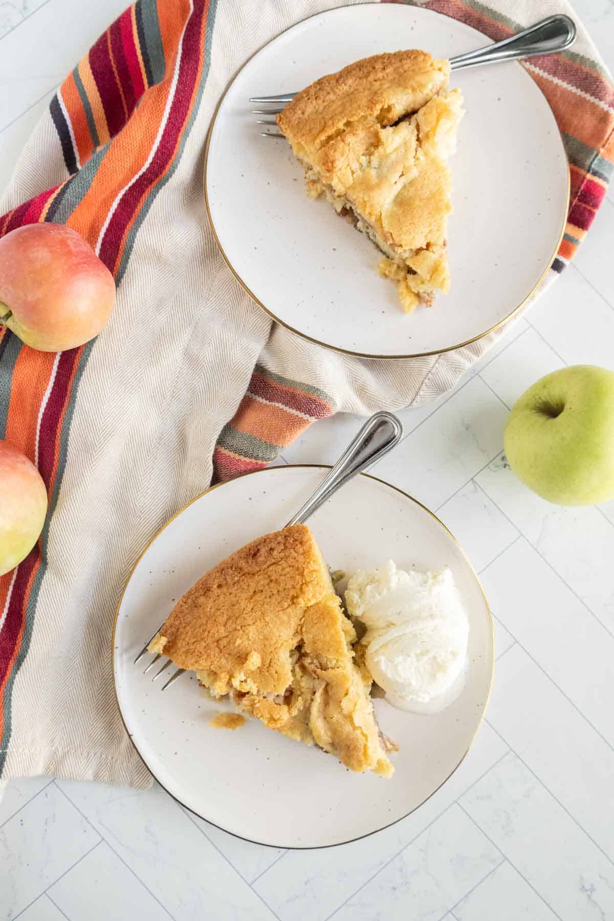 Two plates with apple pie slices, each with a fork, placed on a striped cloth. One plate includes a scoop of vanilla ice cream. Apples are scattered nearby.