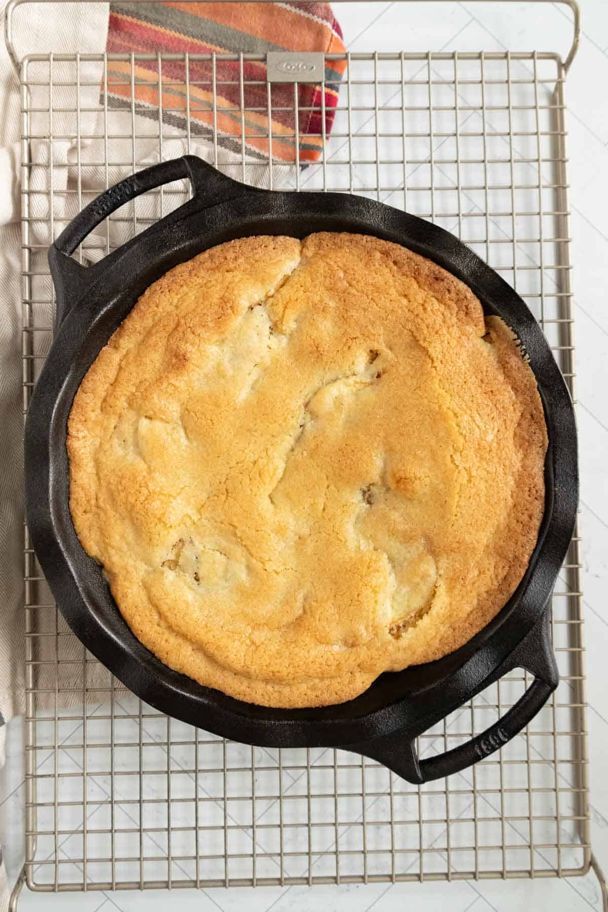 A freshly baked cornbread sits in a cast iron skillet atop a cooling rack. A dish towel with striped edges is partially visible.