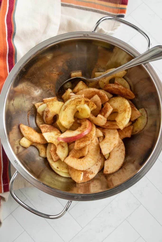 Sliced apples seasoned with cinnamon in a large metal bowl, with a spoon resting inside.