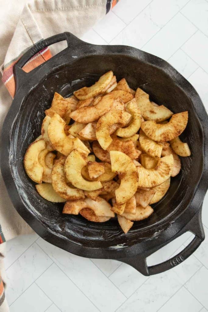 Sliced apples coated with cinnamon in a black cast iron pan, placed on a table with a cloth underneath.