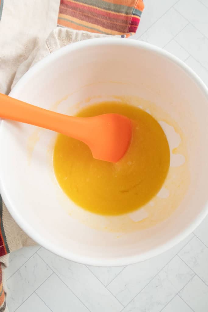 A white bowl with orange liquid mixture and an orange spatula on a light surface.