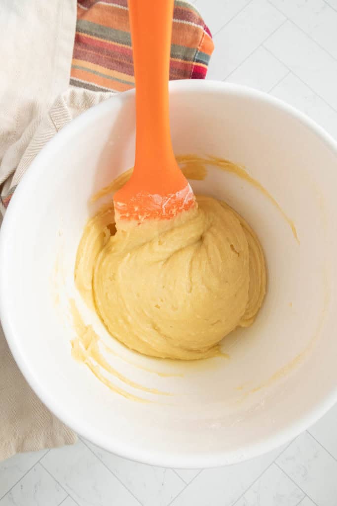 A white bowl with yellow dough and an orange spatula inside, resting on a striped cloth.