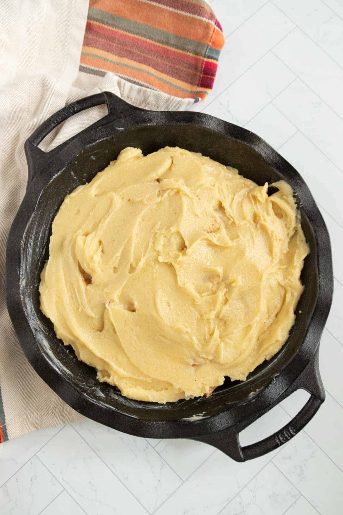 Cast iron skillet with unbaked dough, set on a white countertop with patterned cloth in the background.