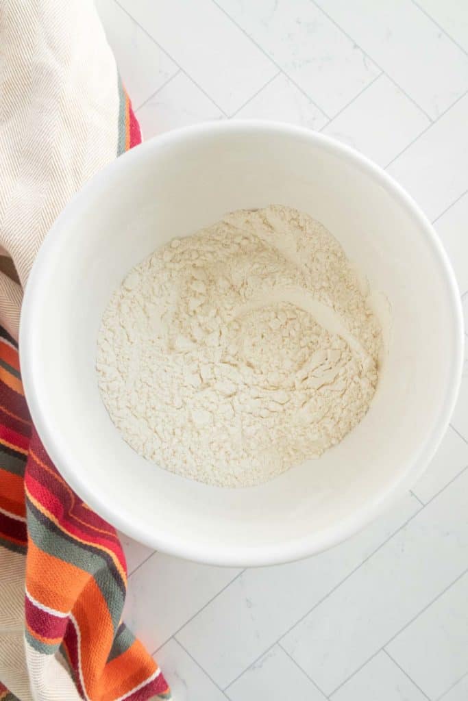 A white bowl filled with flour on a tiled surface, next to a colorful striped towel.