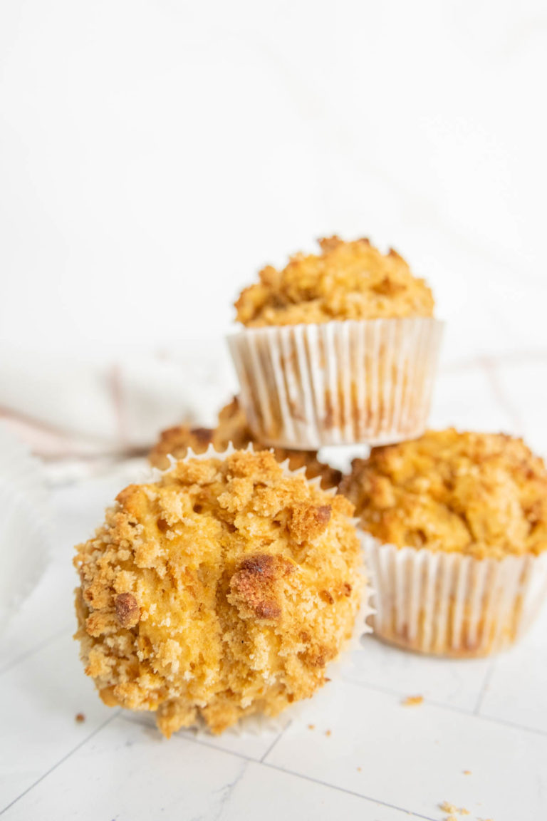 Three muffins with crumbly tops are placed on a white surface, with one muffin in the foreground and two stacked in the background.