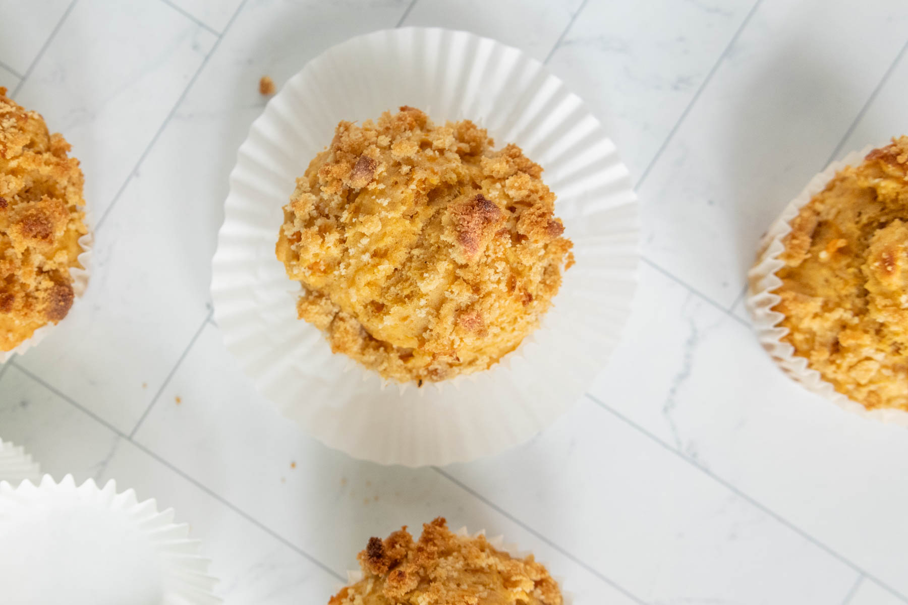 Muffins with crumbly tops on white paper liners laid out on a white tiled surface.