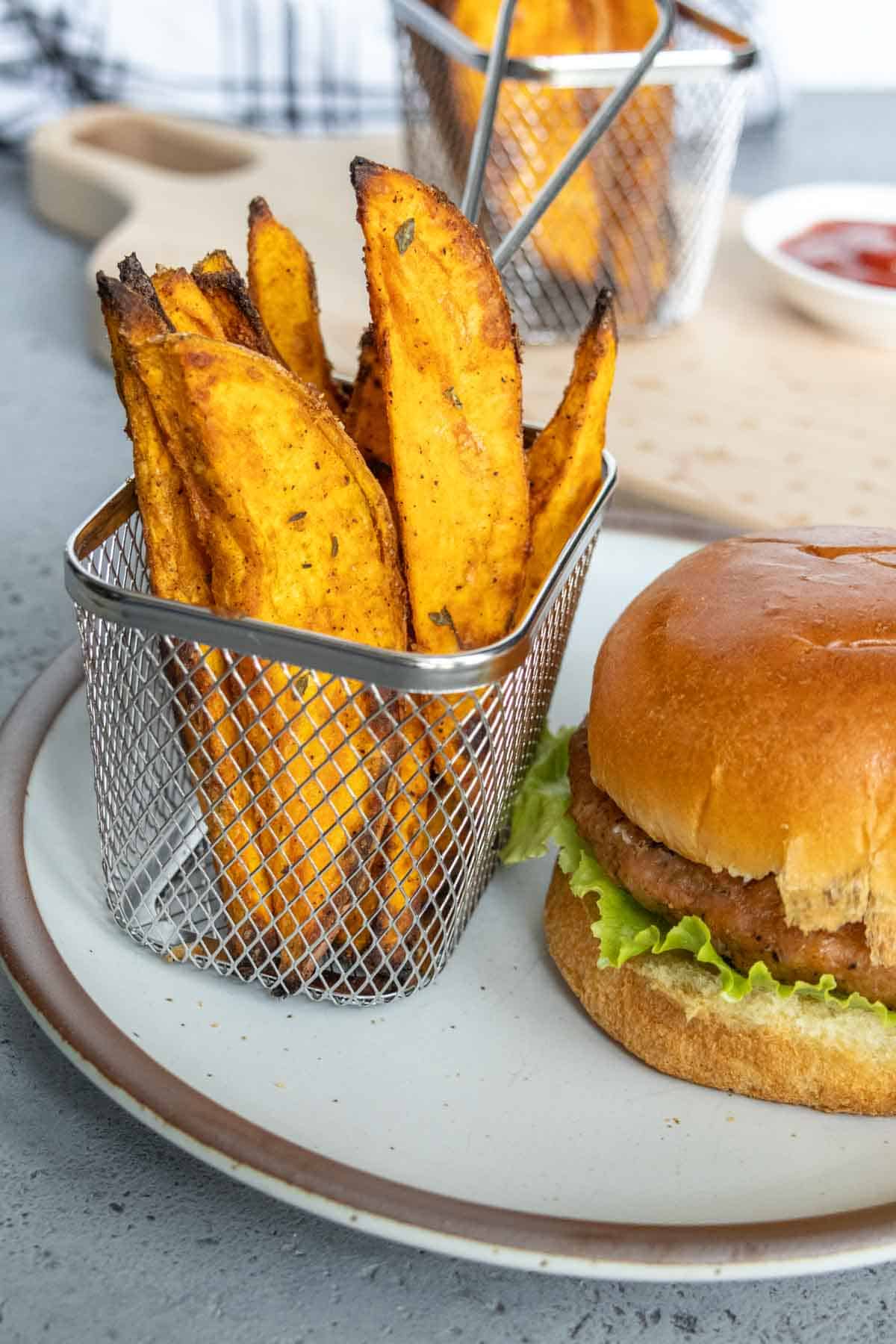 A plate with a burger in a bun with lettuce and a metal basket of seasoned sweet potato fries, and a dip in the background.