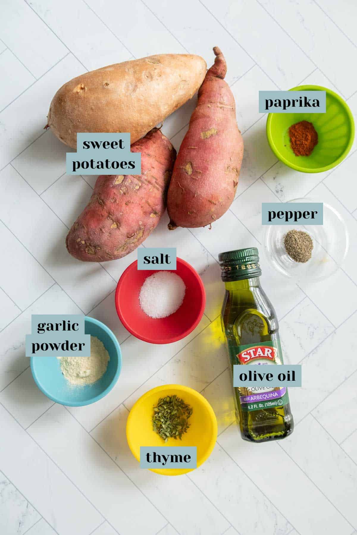 Ingredients for a recipe including sweet potatoes, garlic powder, salt, thyme, olive oil, paprika, and pepper arranged on a white surface. Each ingredient is labeled.