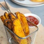 Basket of sweet potato fries served with a small dish of ketchup on a wooden board. A burger on a plate is slightly blurred in the background.