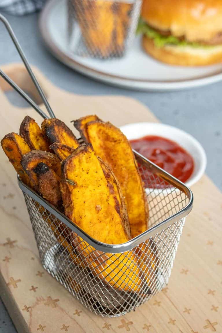 Basket of sweet potato fries served with a small dish of ketchup on a wooden board. A burger on a plate is slightly blurred in the background.