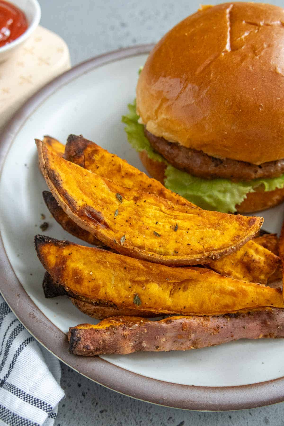 A plate with a sandwich in a bun containing a meat patty and lettuce. Next to it, there are three seasoned sweet potato wedges. A small part of a napkin and a ketchup dish are visible in the background.