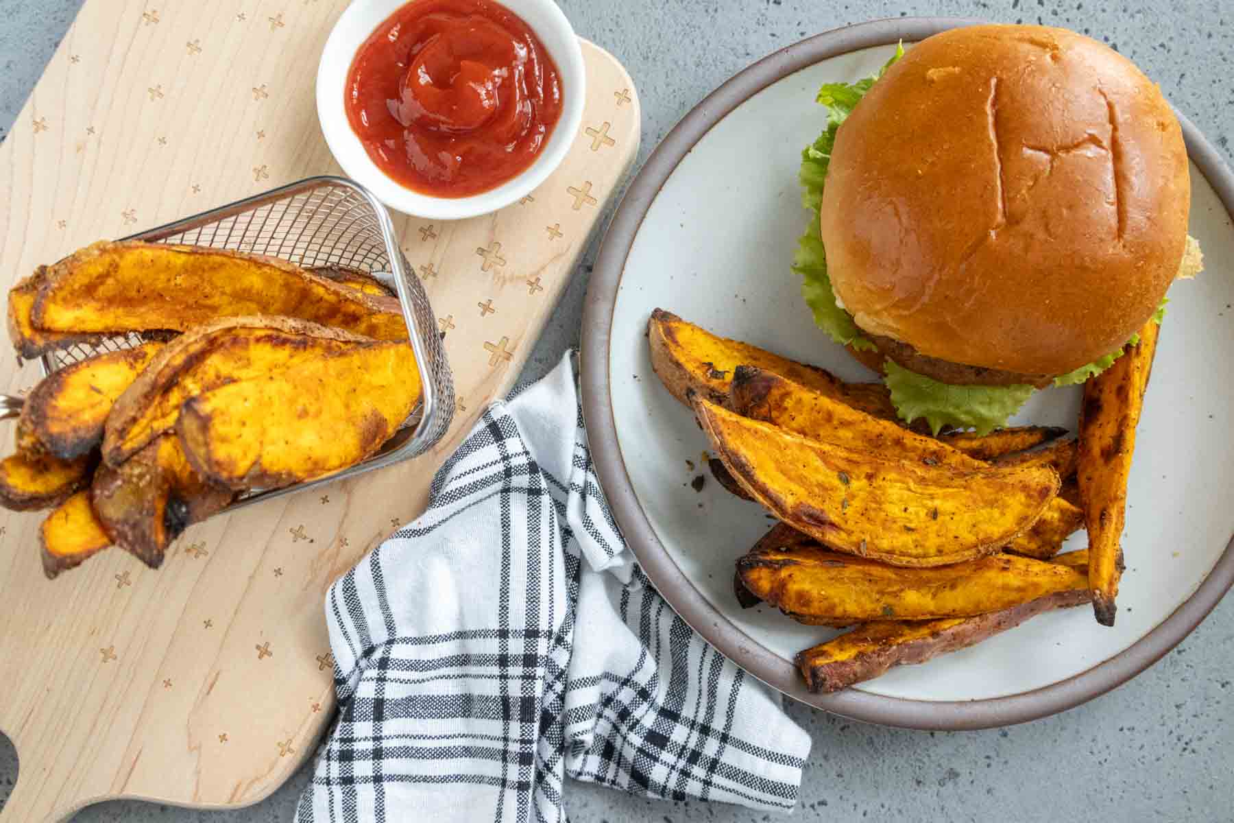 A plate with a burger and sweet potato wedges, a small basket of sweet potato wedges, a bowl of ketchup, and a black-and-white striped napkin on a wooden board.