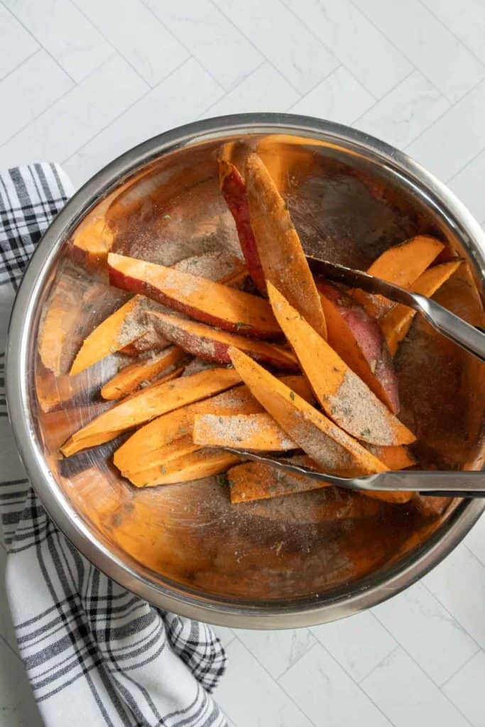 A stainless steel bowl filled with raw, sliced sweet potatoes, seasoned with spices, and being mixed with metal tongs. A checkered cloth is partially visible on the side.