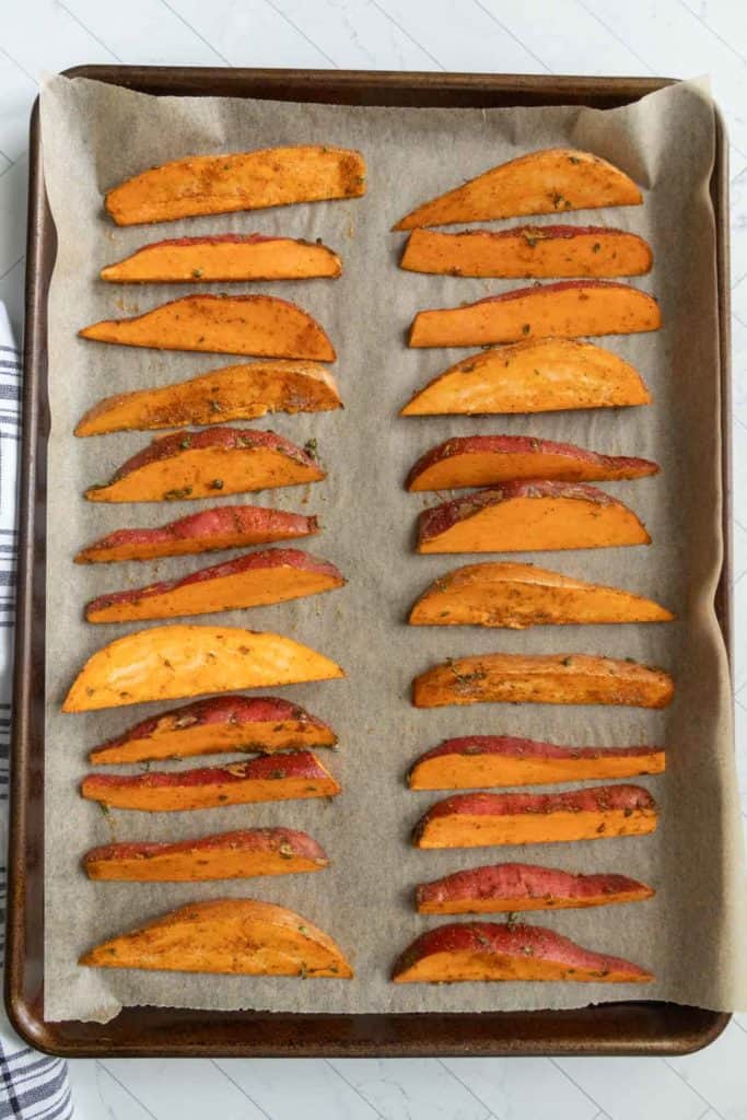 A baking tray lined with parchment paper holds evenly spaced, seasoned sweet potato wedges arranged in rows.