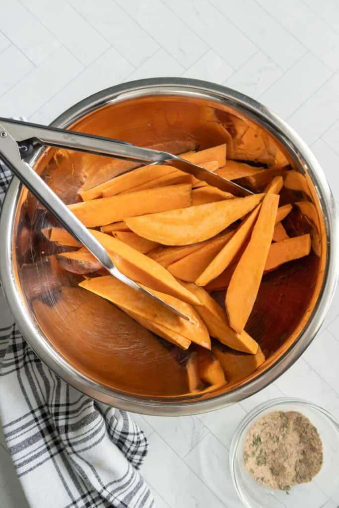 Metal bowl with raw sweet potato wedges and tongs set on a white surface next to a small bowl containing seasoning.