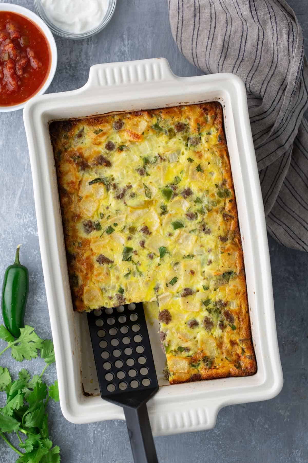 A baked casserole with a serving spatula, featuring visible ingredients like ground meat, cheese, and vegetables. Nearby are a jalapeño, cilantro, and bowls of salsa and sour cream.