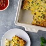 Baked casserole with a slice on a plate, accompanied by a fork. Salsa and sour cream in bowls are nearby. Parsley is placed at the bottom right corner.