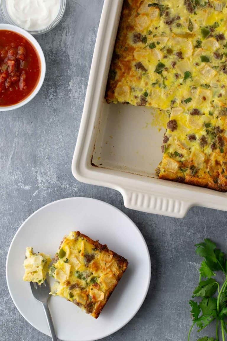 Baked casserole with a slice on a plate, accompanied by a fork. Salsa and sour cream in bowls are nearby. Parsley is placed at the bottom right corner.