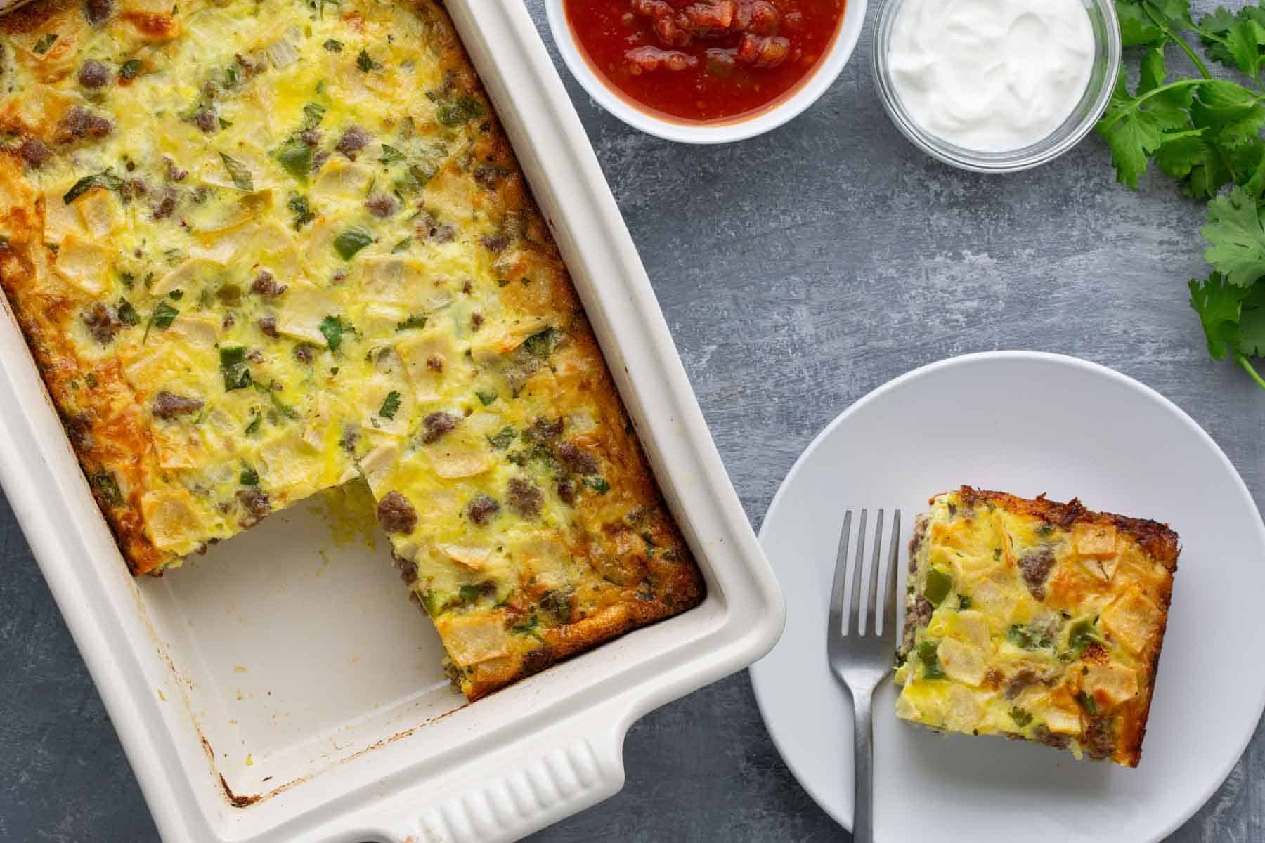 Baked casserole with a cut slice on a white plate, accompanied by salsa and sour cream in small bowls. A sprig of cilantro is placed nearby.