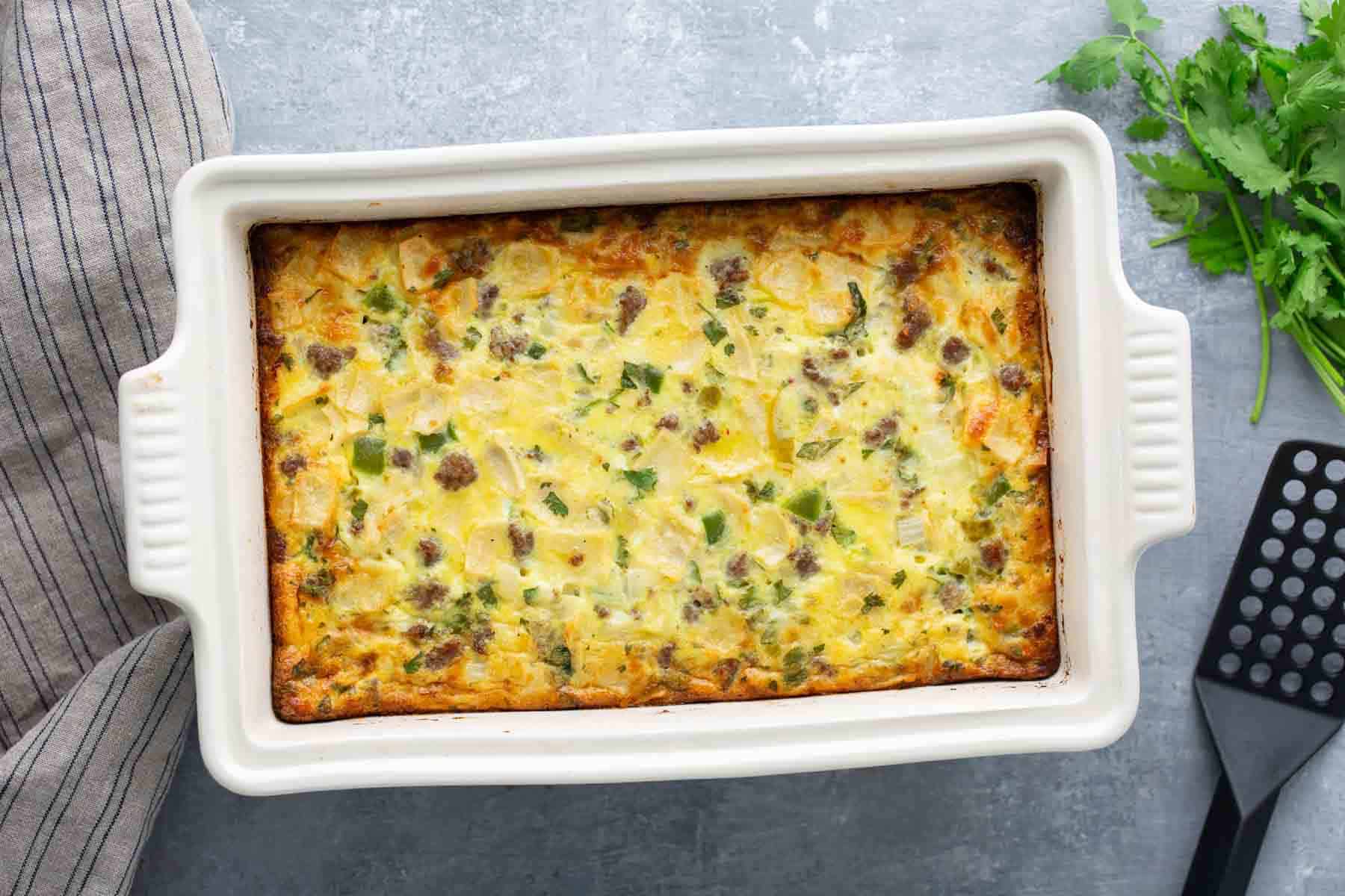 A baked casserole with eggs, sausage, cheese, and vegetables in a white rectangular dish. A spatula and parsley are on the side.