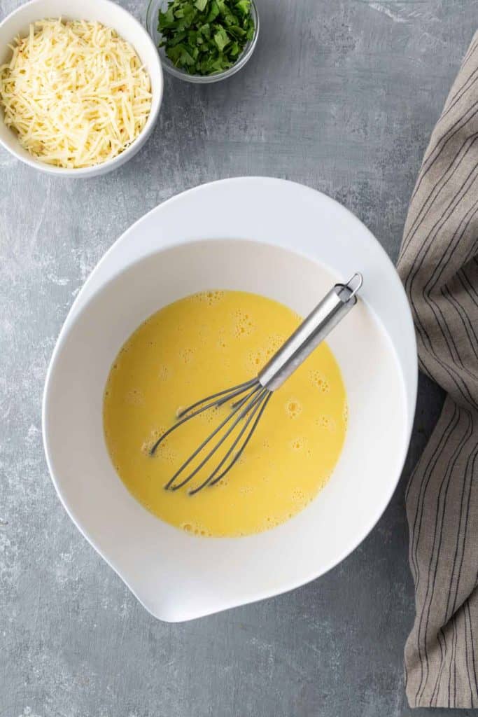 A white bowl with beaten eggs and a whisk, surrounded by grated cheese and chopped herbs in small bowls on a gray surface.