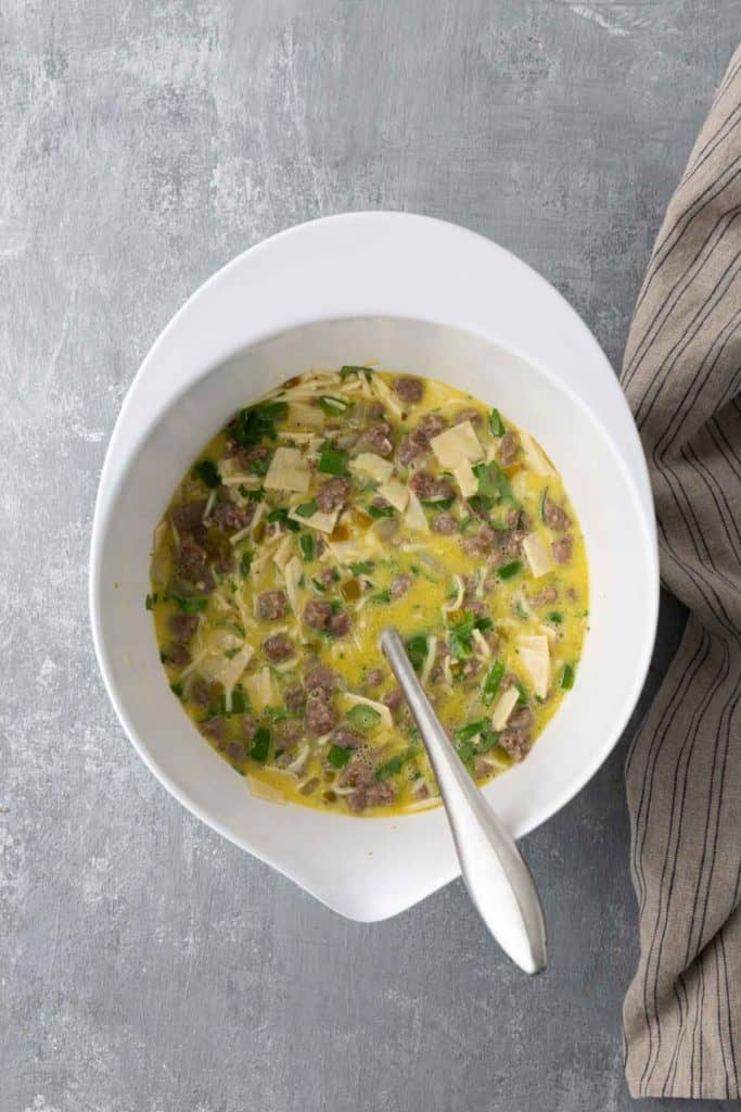 A bowl of soup with pieces of meat, noodles, and green onions, accompanied by a silver spoon. A striped cloth is partially visible on the side.