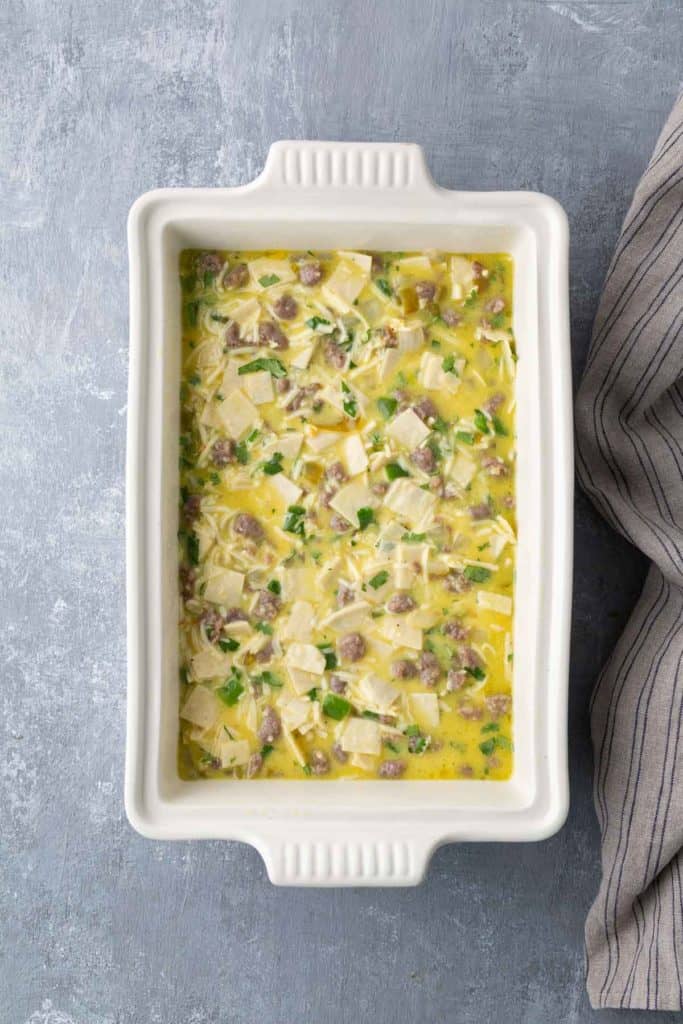 Rectangular baking dish filled with uncooked breakfast casserole, featuring eggs, sausage, and cheese, on a textured gray surface.