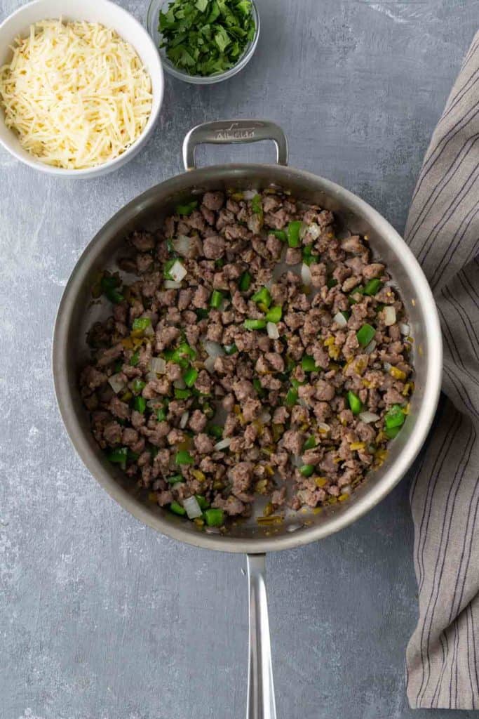 A skillet with browned ground beef, chopped green peppers, and onions. Next to it are bowls of shredded cheese and chopped herbs.