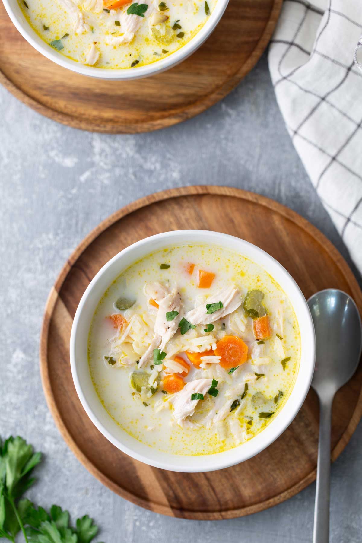 A bowl of chicken and vegetable soup with orzo, garnished with parsley on a wooden coaster. A spoon is placed beside the bowl.