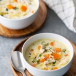 Two bowls of creamy chicken soup with carrots and herbs on wooden trays. A spoon is placed next to one bowl, and a checkered cloth is in the background.